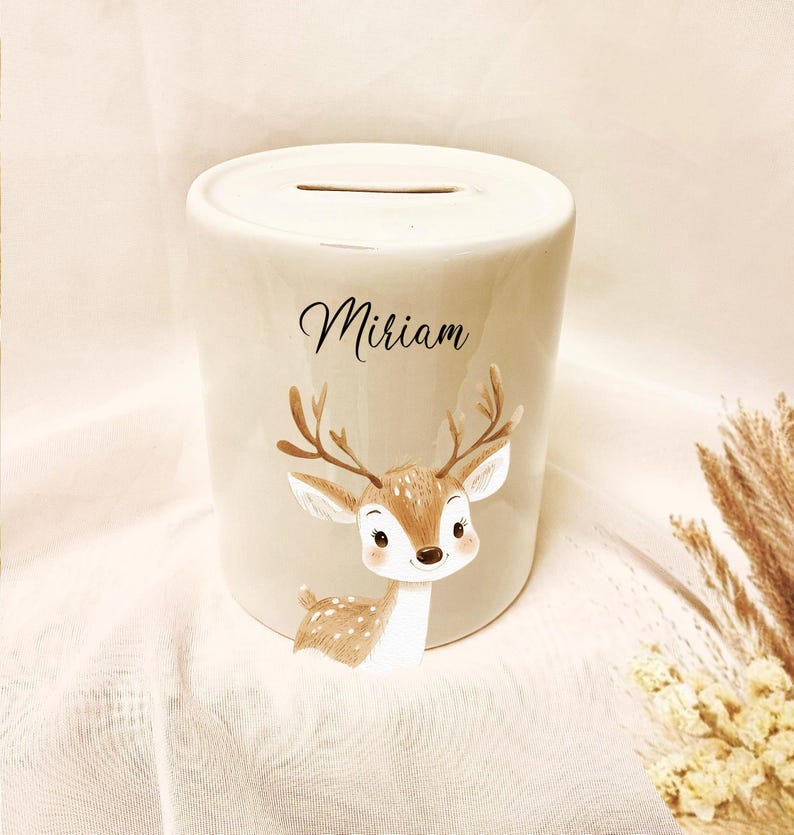 May include: A cream-colored ceramic bank with a slot on top. The bank features a cartoon deer illustration in brown and white, with the name "Miriam" in black script. The bank is cylindrical and sits on a white surface.