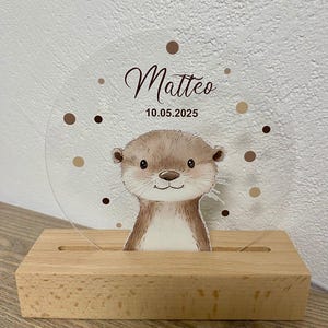 K&ouml;nnte beinhalten: Transparente Acrylplatte mit einer Cartoon-Otter-Illustration, dem Namen "Matteo" in Schreibschrift und dem Datum 10.05.2025. Die Platte steht in einem Holzsockel. Braune Punkte umgeben den Namen und das Datum.