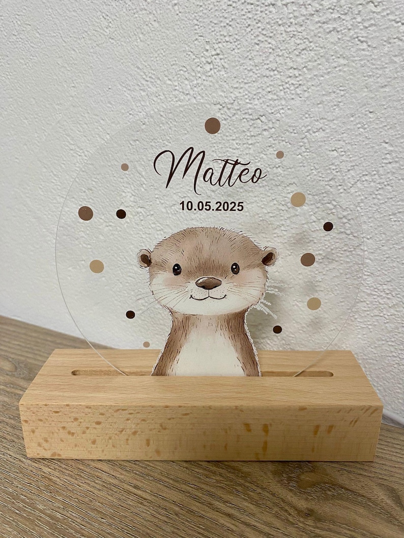 K&ouml;nnte beinhalten: Transparente Acrylplatte mit einer Cartoon-Otter-Illustration, dem Namen "Matteo" in Schreibschrift und dem Datum 10.05.2025. Die Platte steht in einem Holzsockel. Braune Punkte umgeben den Namen und das Datum.