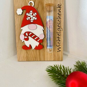 May include: Wooden Christmas gnome ornament with a red hat, white beard, and candy cane. A glass tube with a cork top is attached, containing rolled-up paper money. The text "Wichtelgeschenk" is etched on the wood.