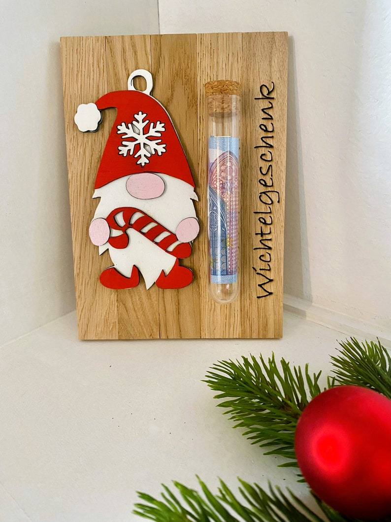 May include: Wooden Christmas gnome ornament with a red hat, white beard, and candy cane. A glass tube with a cork top is attached, containing rolled-up paper money. The text "Wichtelgeschenk" is etched on the wood.