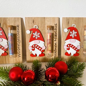 May include: Wooden Christmas gnome ornaments with red hats, white beards, and candy cane accents. Each ornament holds a glass tube with money inside. The text on the wood reads "Wichtelgeschenk", "Sch&ouml;nen Nikolaus", and "Frohe Weihnachten".
