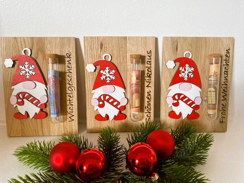 May include: Wooden Christmas gnome ornaments with red hats, white beards, and candy cane accents. Each ornament holds a glass tube with money inside. The text on the wood reads "Wichtelgeschenk", "Sch&ouml;nen Nikolaus", and "Frohe Weihnachten".