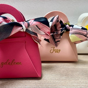 Op de afbeelding: Kleine, handtasvormige geschenkdozen in roze, perzik en wit. Elke doos heeft een handvat en een sjaal met patroon. De roze doos heeft de naam "Magdalena" in gouden letters.