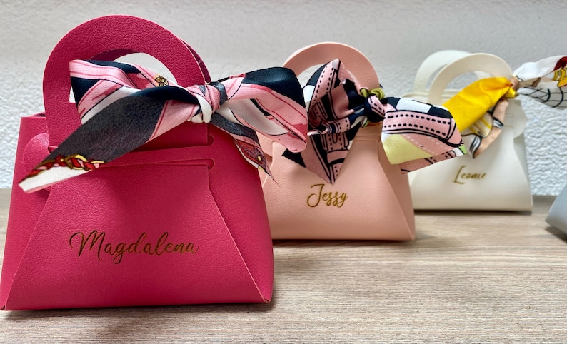 Op de afbeelding: Kleine, handtasvormige geschenkdozen in roze, perzik en wit. Elke doos heeft een handvat en een sjaal met patroon. De roze doos heeft de naam "Magdalena" in gouden letters.