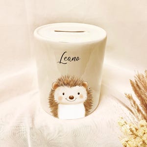 May include: A white ceramic piggy bank with a slot on top. The bank features a cartoon hedgehog illustration with a smiling face and the name "Leano" in script. The hedgehog has brown quills and a white belly.