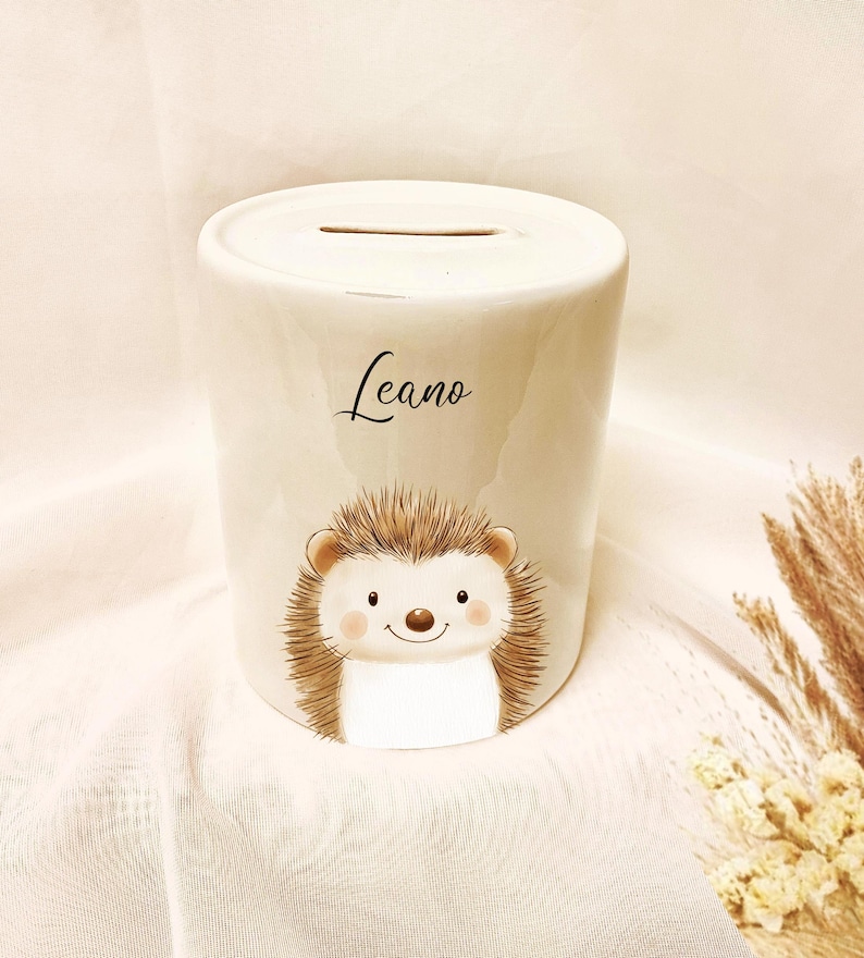 May include: A white ceramic piggy bank with a slot on top. The bank features a cartoon hedgehog illustration with a smiling face and the name "Leano" in script. The hedgehog has brown quills and a white belly.
