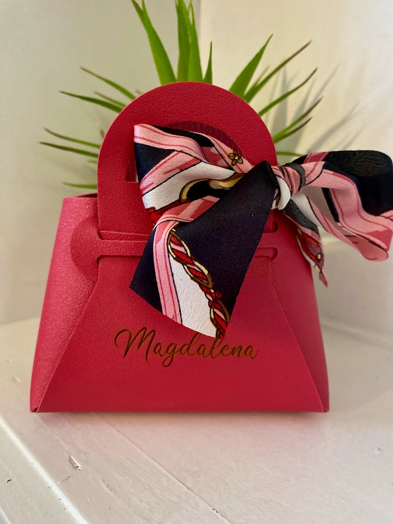 Op de afbeelding: Een felroze cadeauzakje in de vorm van een handtas, met een handvat en een decoratieve sjaal eraan vastgebonden. De sjaal heeft een patroon van roze, zwart en goud. De naam "Magdalena" staat op de voorkant van de tas.