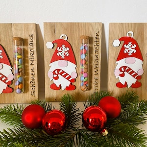 May include: Wooden Christmas decorations featuring gnome figures with red hats and candy canes, holding tubes filled with colorful candies. The wooden plaques have text in German. Red ornaments and pine branches complete the festive scene.