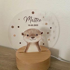 K&ouml;nnte beinhalten: Nachtlicht mit Holzsockel und Otter-Illustration. Das klare Acryllicht tr&auml;gt den Namen "Matteo" und das Datum "10.05.2025", umgeben von braunen Punkten. Der Sockel besteht aus hellem Holz.