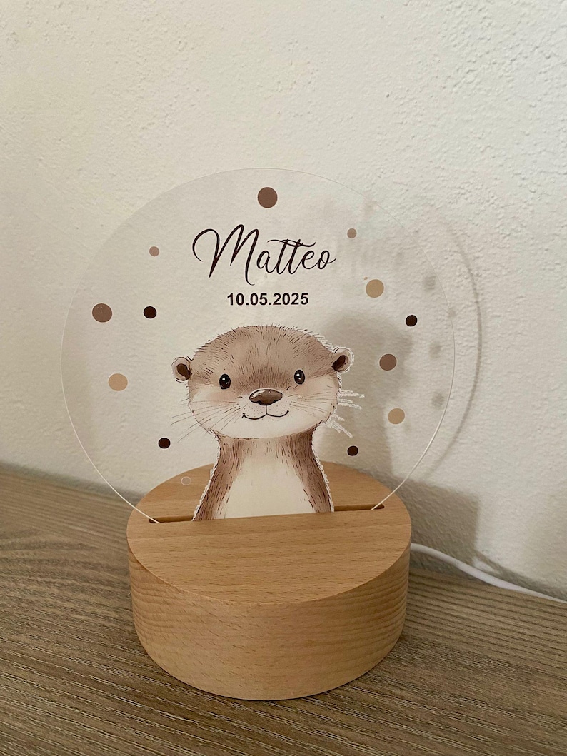 K&ouml;nnte beinhalten: Nachtlicht mit Holzsockel und Otter-Illustration. Das klare Acryllicht tr&auml;gt den Namen "Matteo" und das Datum "10.05.2025", umgeben von braunen Punkten. Der Sockel besteht aus hellem Holz.