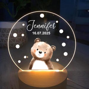 K&ouml;nnte beinhalten: Personalisiertes Nachtlicht mit einer B&auml;ren-Illustration. Die transparente Acrylplatte wird von einem warmen Licht beleuchtet, mit dem Namen "Jennifer" und dem Datum "16.07.2025". Der Sockel ist aus Holz.
