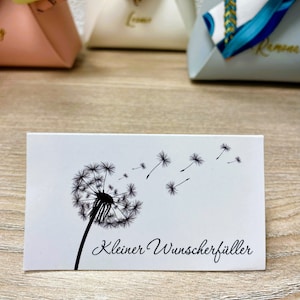 Op de afbeelding: Een witte rechthoekige kaart met een zwarte paardenbloemillustratie en wegvliegende zaden. De tekst "Kleiner Wunscherf&uuml;ller" staat in script eronder. De kaart ligt op een houten oppervlak.