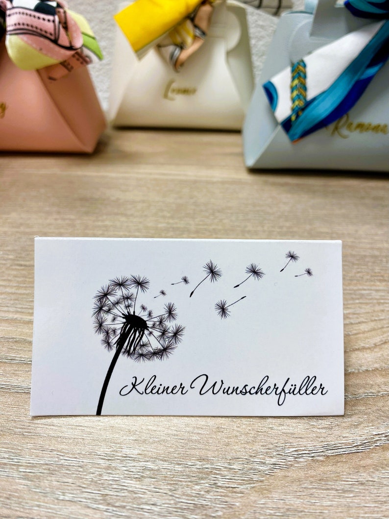 Op de afbeelding: Een witte rechthoekige kaart met een zwarte paardenbloemillustratie en wegvliegende zaden. De tekst "Kleiner Wunscherf&uuml;ller" staat in script eronder. De kaart ligt op een houten oppervlak.