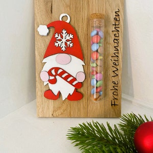 May include: Wooden Christmas decoration featuring a gnome with a red hat and candy cane, alongside a glass tube filled with colorful candies. The words "Frohe Weihnachten" are etched on the wood.