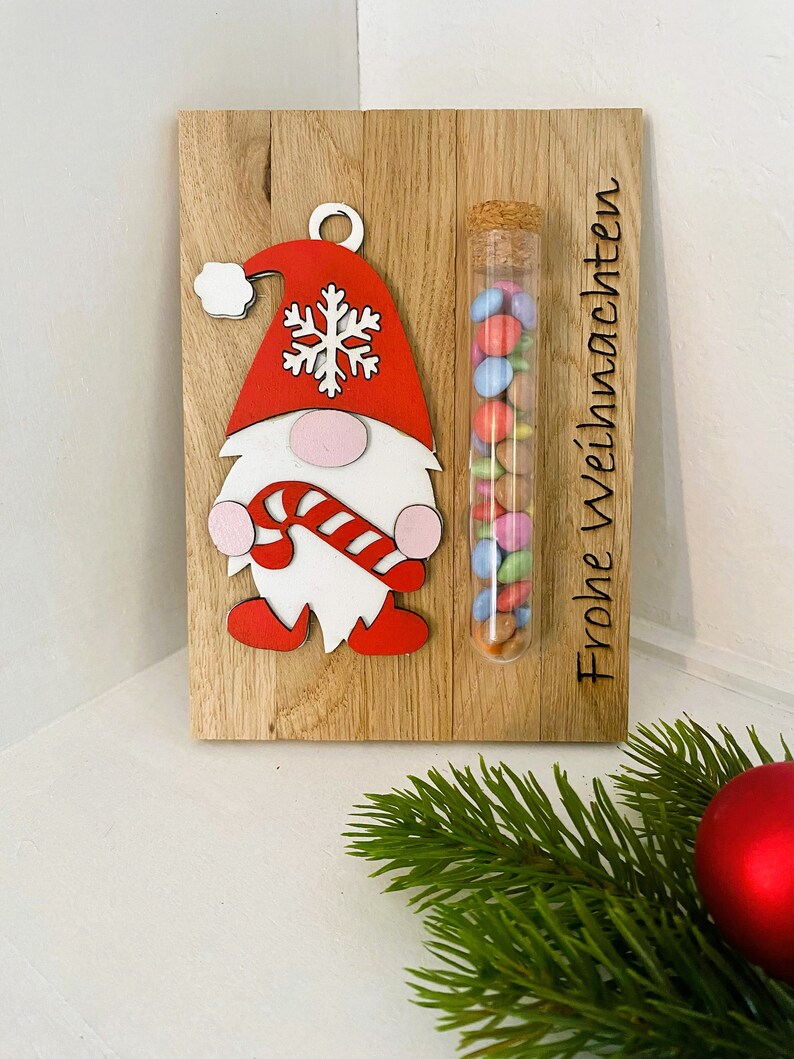 May include: Wooden Christmas decoration featuring a gnome with a red hat and candy cane, alongside a glass tube filled with colorful candies. The words "Frohe Weihnachten" are etched on the wood.