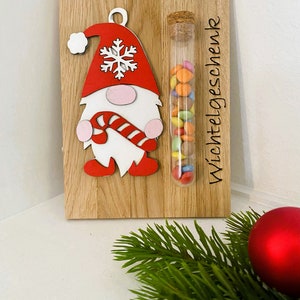 May include: Wooden Christmas decoration featuring a gnome with a red hat and candy cane. A glass tube filled with colorful candies is attached to the wooden board. The text "Wichtelgeschenk" is printed on the board.