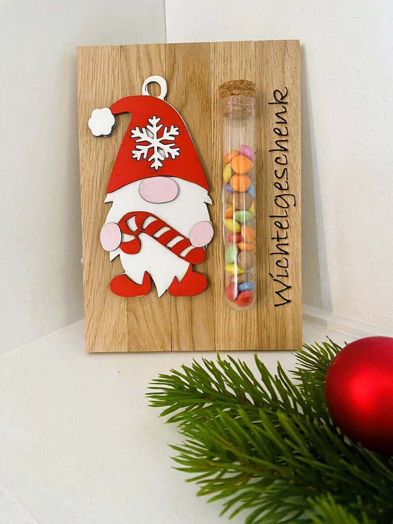 May include: Wooden Christmas decoration featuring a gnome with a red hat and candy cane. A glass tube filled with colorful candies is attached to the wooden board. The text "Wichtelgeschenk" is printed on the board.