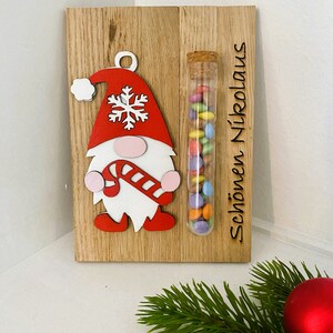 May include: Wooden Christmas decoration featuring a gnome with a red hat and candy cane. A glass tube filled with colorful candies is attached. The text "Sch&ouml;nen Nikolaus" is engraved on the wood.