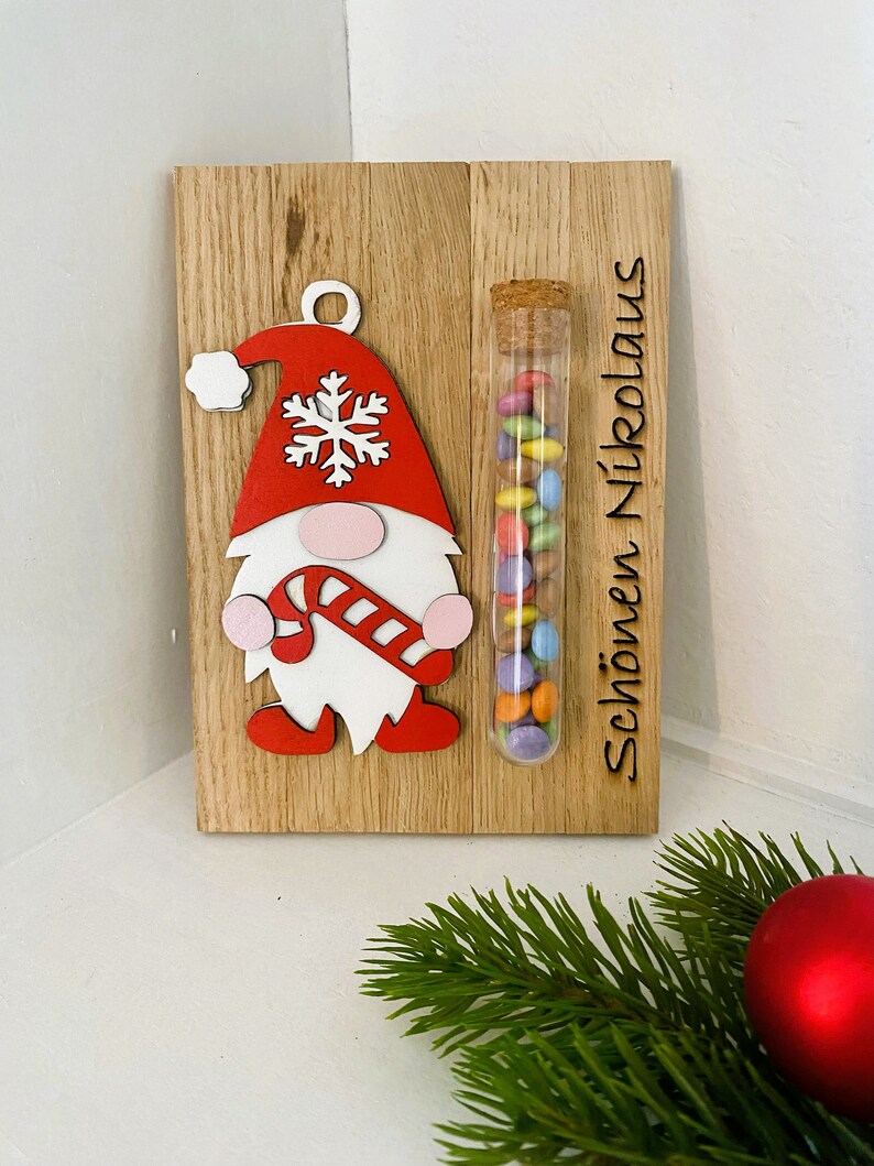May include: Wooden Christmas decoration featuring a gnome with a red hat and candy cane. A glass tube filled with colorful candies is attached. The text "Sch&ouml;nen Nikolaus" is engraved on the wood.