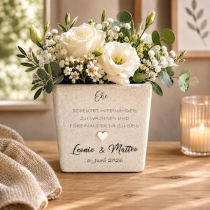 Könnte beinhalten: Ein quadratischer, strukturierter, hellfarbener Pflanztopf gefüllt mit weißen Rosen, kleinen weißen Blüten und Grünzeug. Der Pflanztopf trägt den Text "Ehe bedeutet, miteinander zu wachsen und füreinander da zu sein." Darunter ein Herz und die Namen "Leonie & Matteo" und das Datum "6. Juni 2026."