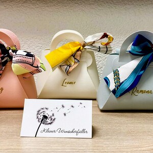 Op de afbeelding: Een verzameling kleurrijke cadeauzakjes, elk lijkend op een handtas, met een handvat en een decoratieve sjaal die aan de bovenkant is vastgebonden. De zakjes zijn in roze, perzik, cr&egrave;me, lichtblauw en marineblauw. Elke tas heeft een naam.