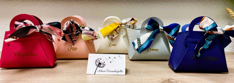 Op de afbeelding: Een verzameling kleurrijke cadeauzakjes, elk lijkend op een handtas, met een handvat en een decoratieve sjaal die aan de bovenkant is vastgebonden. De zakjes zijn in roze, perzik, cr&egrave;me, lichtblauw en marineblauw. Elke tas heeft een naam.