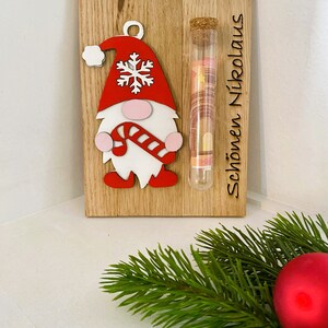 May include: Wooden Christmas decoration featuring a gnome with a red hat and candy cane, a glass tube with money, and the text "Sch&ouml;nen Nikolaus". The gnome has a white beard and pink cheeks. The background is a light wood.