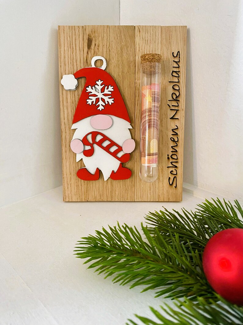 May include: Wooden Christmas decoration featuring a gnome with a red hat and candy cane, a glass tube with money, and the text "Sch&ouml;nen Nikolaus". The gnome has a white beard and pink cheeks. The background is a light wood.