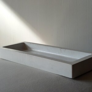 Può includere: Un vassoio rettangolare in cemento grigio chiaro con un interno poco profondo. Il vassoio ha un design semplice e minimalista con linee pulite e una superficie liscia. Il vassoio è adatto per organizzare piccoli oggetti.