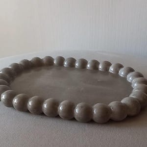 Può includere: Un vassoio decorativo ovale grigio con un bordo a perline. Le perline sono di un colore grigio tenue e sono uniformemente distanziate attorno al perimetro del vassoio. Il vassoio è su una superficie grigio chiaro.