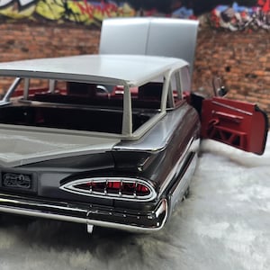 Puede incluir: Un detallado modelo de coche en negro y plata con techo blanco y maletero abierto. La puerta roja está abierta, revelando el interior. El coche tiene detalles cromados y un diseño clásico, sobre un fondo de pared de ladrillo.