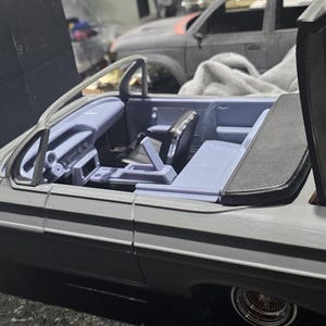 Könnte beinhalten: Ein graues Modell eines Cabriolets mit geöffneter Motorhaube und Türen, das einen detaillierten Innenraum mit blauen Sitzen und Armaturenbrett zeigt. Das Auto hat goldfarbene Akzente und ist vor einem verschwommenen Hintergrund platziert.