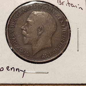 May include: A vintage British penny coin, featuring a profile of King George V, is displayed in a protective holder. The coin's inscription includes "GEORGIVS V DEI GRA BRITT OMN REX FID DEF IND IMP". The holder is labeled "1 penny" and "Britain".
