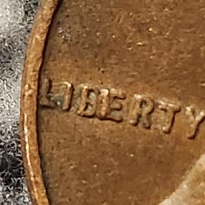 May include: Close-up of a copper-colored coin with the word "LIBERTY" embossed on its surface. The coin's edge and surface texture are visible, suggesting it is an older coin.