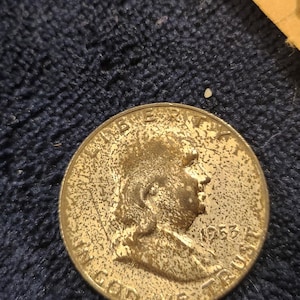 May include: A gold-colored coin featuring a profile of a man and the year 1953. The coin is slightly worn and has a textured surface. The coin is likely a collectible or historical item.