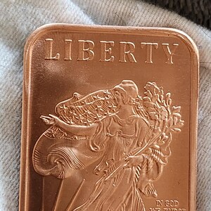 May include: A copper rectangular bar with the word "LIBERTY" at the top. The bar features a detailed relief of a woman in flowing robes, holding a branch. The text "IN GOD WE TRUST" and "ONE OZ .999 FINE COPPER" are also visible.