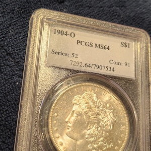 May include: A silver 1904-O Morgan silver dollar coin, graded PCGS MS64, encased in a clear protective holder. The coin features a profile of a woman and the text "1904". The holder includes the text "S$1".