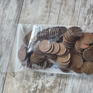 May include: A clear plastic bag filled with numerous copper-colored pennies. The coins are stacked and scattered, with the word "ONE" visible on some. The bag is resting on a light-colored wooden surface.