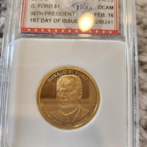 May include: A gold-colored coin with a portrait of Gerald R. Ford, the 38th President of the United States, and the inscription "GERALD R. FORD 38th PRESIDENT 1974-1977". The coin is in a plastic case with a label that reads "2016 S G. FORD $1 38TH PRESIDENT 1ST DAY OF ISSUE PR-70 DCAM FEB. 16 6286241".