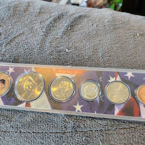 May include: A clear plastic case containing a set of 7 gold-colored coins with a red, white, and blue American flag design. The coins feature various images, including a portrait of a woman and a ship.