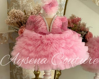 May include: A pink tulle dress with a feathered bodice and a rhinestone embellishment. The dress is on a white stand and has a pink headband with feathers.