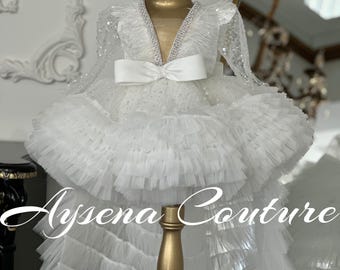 May include: White tulle dress with a high low hemline, featuring a V-neckline, long sleeves, and a satin bow at the waist. The dress is adorned with sequins and rhinestones.