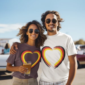 Puede incluir: Dos personas con camisetas con un dise&ntilde;o de coraz&oacute;n en los colores de la bandera alemana. El hombre lleva una camiseta blanca y la mujer una camiseta marr&oacute;n. Ambos sonr&iacute;en y miran a la c&aacute;mara.