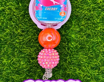 May include: A badge reel featuring an Alani Nu Orange Kiss energy drink can charm, a smooth orange bead, and a textured pink rhinestone bead. Below the reel, a purple sign with white text reads: Please specify which drink/flavor you'd like in the personalization box when ordering.
