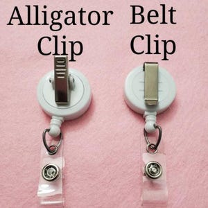 Puede incluir: Dos clips de carrete de insignia blancos con diferentes estilos de clip met&aacute;lico. El clip izquierdo es un clip de cocodrilo, y el clip derecho es un clip de cintur&oacute;n. Ambos tienen una correa de pl&aacute;stico transparente con cierre a presi&oacute;n y un anillo de metal.
