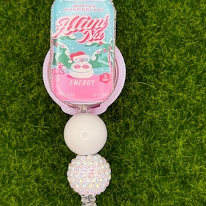 May include: A badge reel featuring a pink and blue can design with the text "Winter Wonderland Alani Nu Energy". The can design has a Santa Claus graphic. The reel has a white and iridescent bead design.