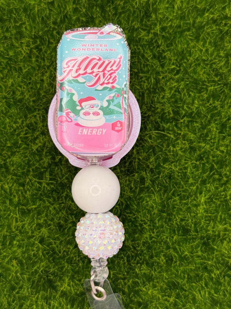 May include: A badge reel featuring a pink and blue can design with the text "Winter Wonderland Alani Nu Energy". The can design has a Santa Claus graphic. The reel has a white and iridescent bead design.