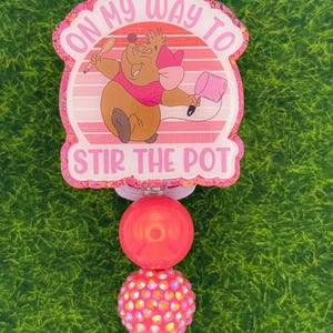 May include: A pink and white badge reel with the text "ON MY WAY TO STIR THE POT" and an illustration of a cartoon mouse. The reel has two pink beads and a clear plastic clip.