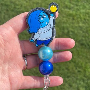 May include: A blue and white glitter badge reel featuring a cartoon character from the movie Inside Out. The character is Sadness, and she is holding a yellow ball. The reel has three beads: one light blue, one dark blue, and one light blue.